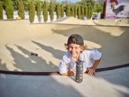 Cooler kleiner Junge mit Schirmmütze hängt an Skaterrampe und trinkt Wasser aus Sportflasche mit Strohhalm, der in die Kamera schaut — Stockfoto