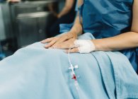 Paciente femenina de cultivo sentada con las piernas cubiertas y aguja de líquido intravenoso en la mano antes de la cirugía en quirófano - foto de stock