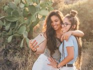 Щасливі щасливі випадкові жінки фотографують на смартфоні, стоячи поруч з кактусом в покинутій сільській місцевості на заході сонця — стокове фото