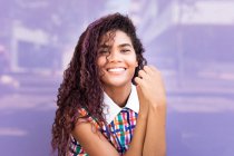 Портрет улыбающейся молодой этнической девушки с вьющимися волосами, смотрящей в камеру на фиолетовой стеклянной стене — стоковое фото