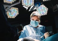 Da sotto grave giovane medico in maschera protettiva e cap facendo chirurgia con strumenti e crop nurse — Foto stock