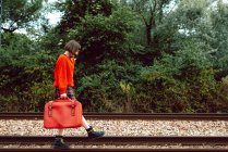 Stilvolle furchtlose Frau mit knallrotem Koffer tritt auf Bahngleise im Grünen — Stockfoto