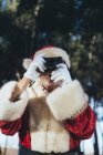 Улыбающийся пожилой человек в костюме Санта-Клауса стоит и фотографирует с камерой на фоне природы — стоковое фото