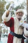 Веселый пожилой человек в костюме Санта-Клауса, стоящий с подарком и колокольчиком в перчатках в руках на фоне природы — стоковое фото