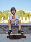 Niño casual con casco y camisa blanca sentado en la rampa en skatepark mirando en la cámara - foto de stock