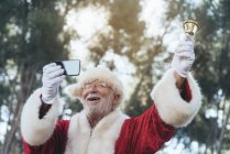 Hombre alegre en traje de Santa Claus timbre campana y tomar selfie con el teléfono móvil en fondo borrosa naturaleza - foto de stock