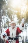Senior man in costume of Santa Claus sitting on cycle, ringing bell and looking at camera — Stock Photo