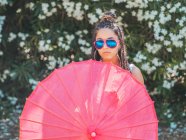 Schlanke junge Frau mit Sonnenbrille und Regenschirm, die neben blühenden Bäumen steht — Stockfoto