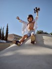 From below of happy boy wearing helmet holding skateboard above head and yelling while sitting on ramp against blue sky — Stock Photo