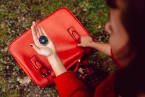Closeup of compass in hand of woman with bright red suitcase on ground with grass — Stock Photo