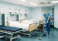 Медик у блакитній формі та захисна маска встановлює лоток на візку в лікарняній кімнаті порожніми ліжками — стокове фото