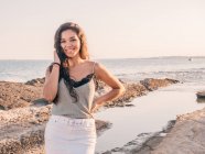 Усміхнений вміст випадкової жінки, що стоїть на пляжі біля берегової лінії на сонячному світлі — стокове фото