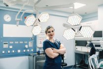 Взрослая женщина в медицинской форме скрещивает руки и смотрит в камеру, стоя возле ламп в современном операционном зале — стоковое фото