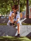 Случайный задумчивый мальчик в наушниках с помощью мобильного телефона, сидящего на скейтборде — стоковое фото