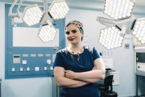 Erwachsene Frau in medizinischer Uniform verschränkt die Arme und blickt in die Kamera, während sie neben Lampen im modernen Operationssaal steht — Stockfoto