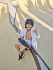 Sonriente niño casual con casco y camisa blanca sentado en la rampa en skatepark mirando en la cámara haciendo un gesto de mano - foto de stock