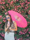 Усміхнена струнка молода жінка в літньому вбранні і сонцезахисних окулярах з парасолькою, що п'є напій біля квітучих дерев — стокове фото