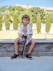 Smiling casual kid in helmet and white shirt sitting on ramp in skatepark looking in camera — Stock Photo
