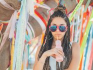 Jeune femme élégante portant des lunettes de soleil boire un cocktail pendant le festival extérieur et regardant la caméra — Photo de stock