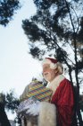 Homme âgé joyeux en costume du Père Noël debout avec des cadeaux et cloche dans les mains gantées sur fond de nature — Photo de stock