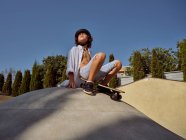 D'en bas de garçon heureux portant un casque tenant skateboard assis sur la rampe contre le ciel bleu — Photo de stock