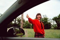 Attractive stylish woman gesturing with thumb up to catch car on road along green field — Stock Photo