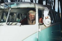 Cheerful senior man in costume of Santa Claus sitting in old van and waving hand from open window — Stock Photo