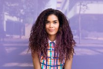 Портрет очаровательной молодой этнической девушки с вьющимися волосами, смотрящей в камеру на фиолетовой стеклянной стене — стоковое фото