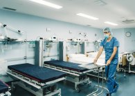 Medic en uniforme azul y bandeja protectora de ajuste de máscara en el carro en la habitación del hospital por camas vacías - foto de stock
