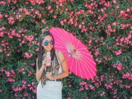 Усміхнена струнка молода жінка в літньому вбранні і сонцезахисних окулярах з парасолькою, що п'є напій біля квітучих дерев — стокове фото