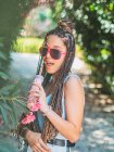 Fröhliche junge Frau mit Sonnenbrille und Dreadlocks trinkt Cocktail und blickt in die Kamera im Park — Stockfoto