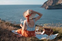 Вид сзади женщины в шляпе с напитком, сидящей на берегу и романтически смотрящей на безмятежную воду — стоковое фото