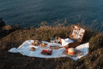 Tappetino da picnic con romantico set con bicchieri di bevande e cibo sulla riva asciutta con acqua serena — Foto stock