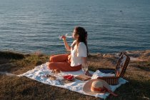 Молода жінка романтично п'є вино на березі біля спокійної води і пагорбів — стокове фото