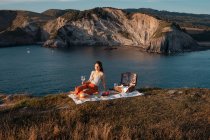 Mujer joven relajándose con bebida en la estera para picnic por el agua serena y colinas - foto de stock