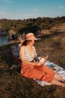 Жінка в капелюсі читає книгу, сидячи на килимку для пікніка на сонячному світлі — стокове фото