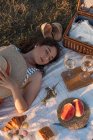 De cima mulher desfrutando deitado no tapete de piquenique branco segurando chapéu cesta nas proximidades no gramado com olhos fechados — Fotografia de Stock