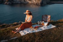 Femme en chapeau lecture livre assis sur le tapis pour pique-nique dans la lumière du soleil — Photo de stock