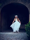 Чарівна маленька дівчинка в повітряній довгій білій сукні та квітковій смузі танцює під аркою в парку — стокове фото