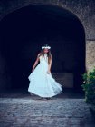 Очаровательная маленькая девочка в белом платье и цветочном повязке, танцующая под аркой в парке — стоковое фото