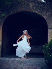 Adorable niña en vestido blanco largo aireado y diadema de flores bailando bajo el arco en el parque mirando a la cámara - foto de stock