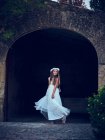 Чарівна маленька дівчинка в повітряній довгій білій сукні та квітковій смузі танцює під аркою в парку, дивлячись далеко — стокове фото