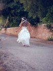 Vista trasera de niña linda irreconocible en vestido blanco aireado y diadema de flores caminando en el parque - foto de stock