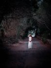 Маленька самотня дівчинка в довгій білій сукні, що стоїть на дорозі в темній алеї, дивлячись далеко — стокове фото