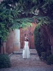 Adorável menina em vestido branco longo arejado e flor headband andando sob arco no parque olhando para longe — Fotografia de Stock