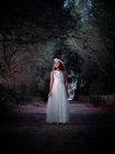 Маленька самотня дівчинка в довгій білій сукні, що стоїть на дорозі в темній алеї, дивлячись далеко — стокове фото