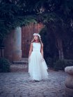 Adorável menina em vestido branco longo arejado e flor headband andando sob arco no parque olhando para longe — Fotografia de Stock