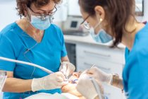 Especialistas seguros en guantes quirúrgicos poniendo sello y trabajando con herramientas dentales en boca del paciente acostado en silla en gabinete - foto de stock