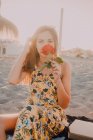 Чарівна вдумлива жінка з трояндою в руці сидить і дивиться в камеру наодинці на сонячному світлі на узбережжі — стокове фото