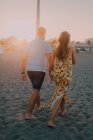 Счастливые влюблённые молодые люди ходят, глядя друг на друга и держась за руки босиком на берегу моря при солнечном свете — стоковое фото